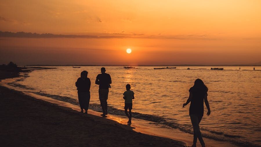 Quatre personnes se promenant sur une plage de sable, avec des bateaux au loin et un coucher de soleil orangé.