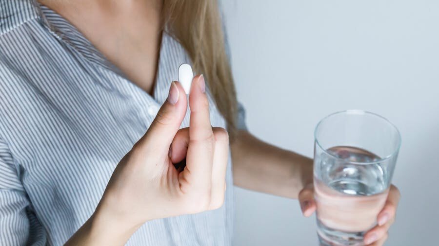 Femme en chemise rayée tenant des médicaments et de l'eau dans un cadre neutre.