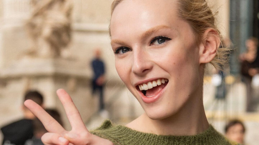 Alex Consani souriante et faisant un signe de paix, vêtue d'un pull rayé vert à la Fashion Week de Paris 2024.