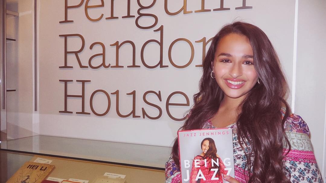 Jazz Jennings souriante tenant son autobiographie « Being Jazz: My Life as a (Transgender) Teen » devant une enseigne Penguin Random House.