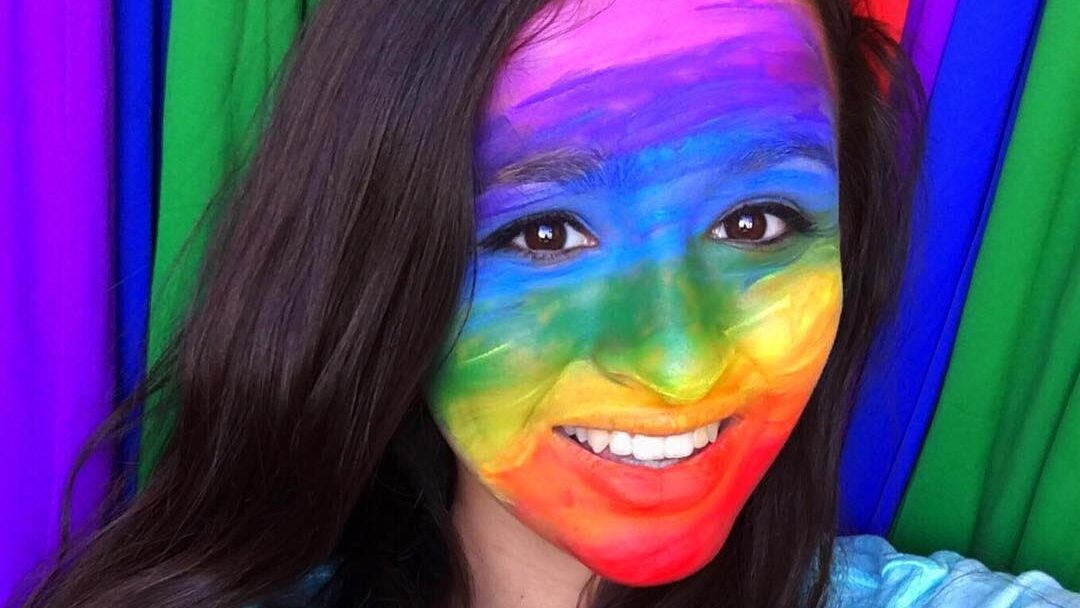 Jazz Jennings souriante devant un fond arc-en-ciel, le visage peint aux couleurs de la fierté.