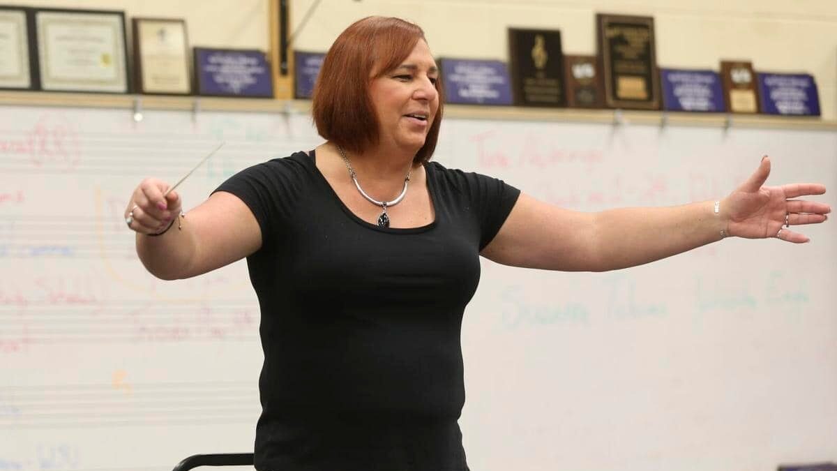 Stephanie Byers devant un tableau blanc, les bras écartés, dirigeant un ensemble musical scolaire dans une salle de classe.