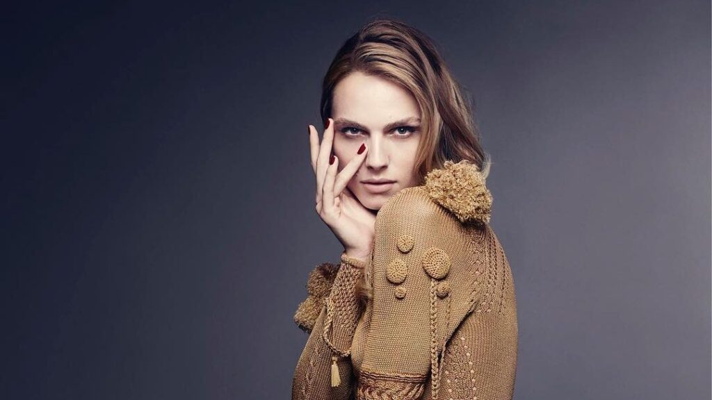 Andreja Pejić portant une robe sculpturale à texture dorée signée Giles Deacon, posant avec grâce dans un portrait éditorial en studio.