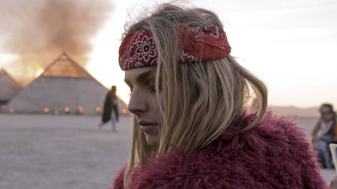 Andreja Pejić dans le désert du Burning Man 2016, bandana rouge et manteau en fausse fourrure, feu derrière elle.