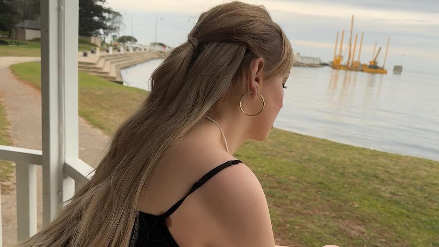 Grace Hyland se tient sur un balcon en bois blanc, penchée en avant en regardant paisiblement l’eau. Ses longs cheveux blonds sont à moitié attachés vers l’arrière. Elle porte un haut noir à fines bretelles et de grandes boucles d’oreilles. La scène est paisible, avec un ciel nuageux et une atmosphère de réflexion.