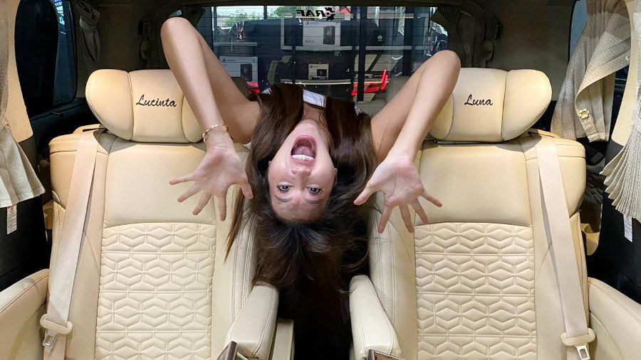 Lucinta Luna suspendue tête en bas entre deux sièges luxueux couleur crème dans son van Alphard personnalisé. Son nom est brodé sur les appuie-têtes et elle fait une expression amusante avec les mains écartées.