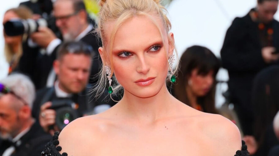 Alex Consani sur un tapis rouge, vêtue d’une robe noire décolletée ornée de perles, coiffure relevée élégante et boucles d’oreilles vertes voyantes.