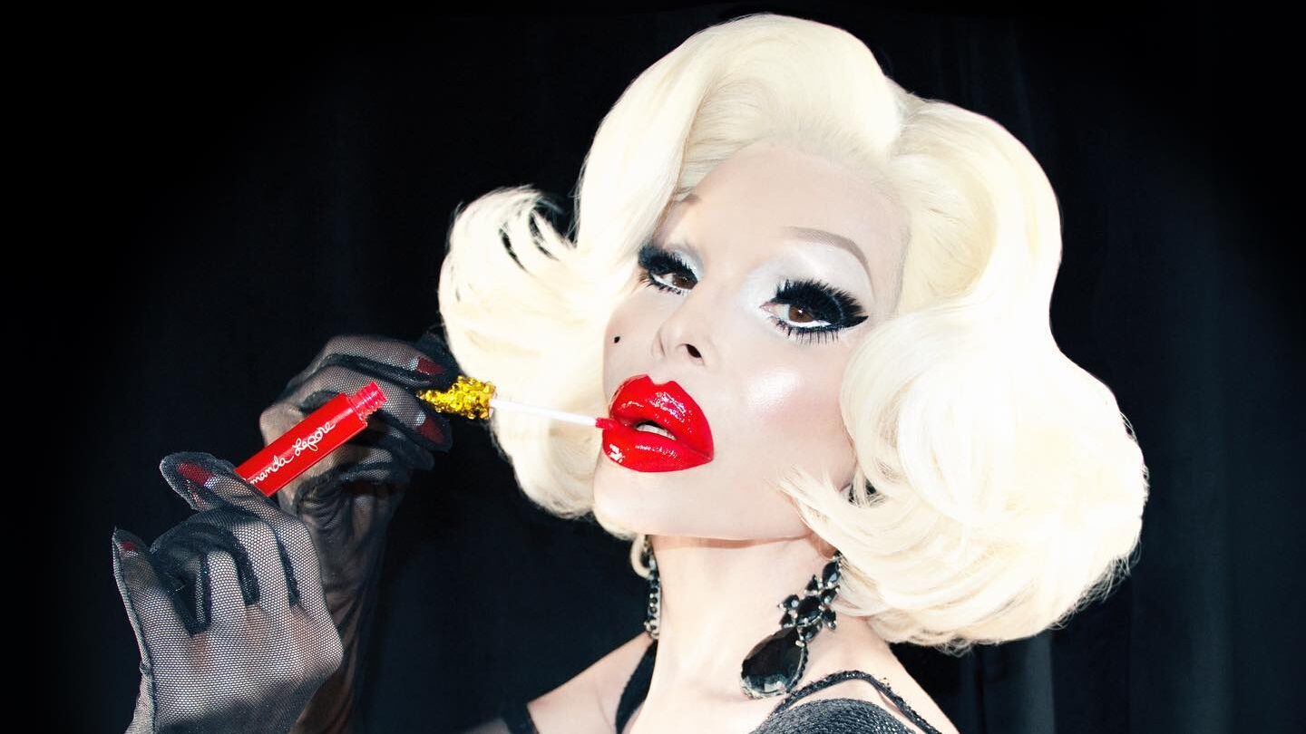 Amanda Lepore aux cheveux blond platine et gloss rouge, tenant l’applicateur devant un fond noir.