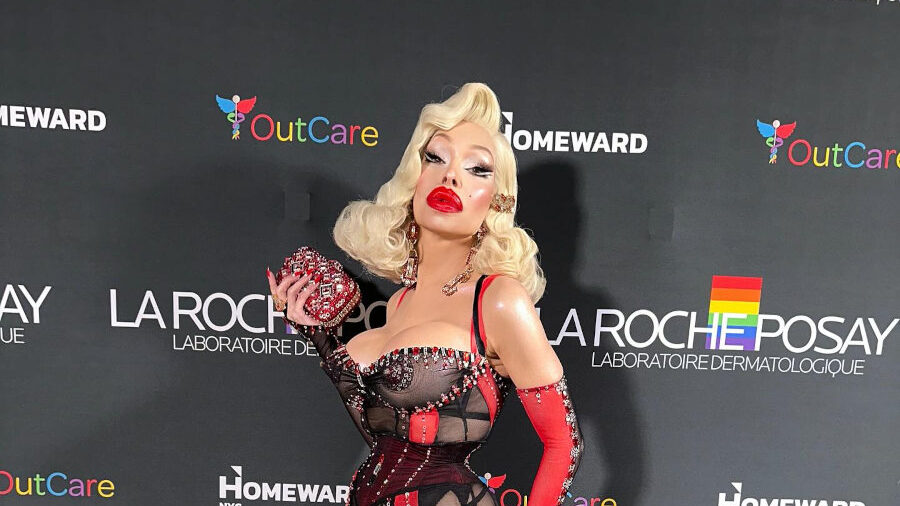 Amanda Lepore posant en tenue noire et rouge sur un tapis arc-en-ciel lors de l’événement Pride de La Roche-Posay, avec logos en arrière-plan.
