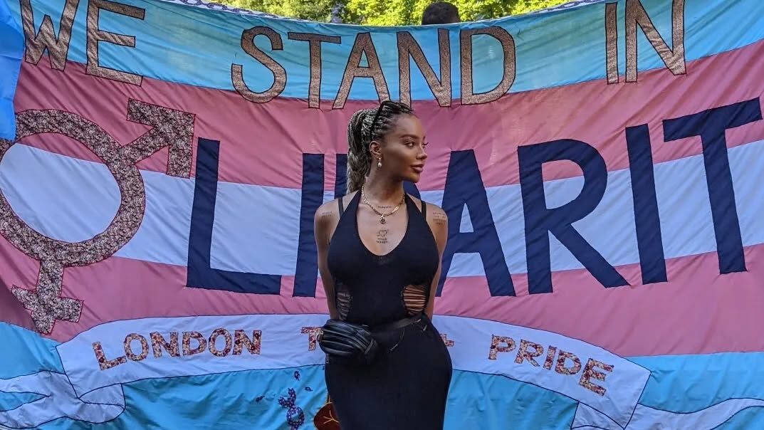 Munroe Bergdorf en longue robe noire posant devant une bannière trans portant l’inscription “We Stand in Solidarity” lors de la Pride 2022.