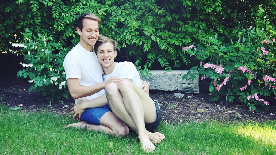 Peter Zurkuhlen et Tommy Dorfman assis sur l’herbe, souriants et enlacés dans un jardin avec des fleurs roses.
