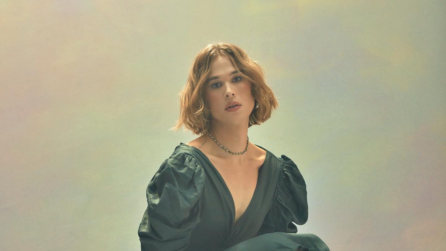 Portrait de Tommy Dorfman en vert, avec cheveux ondulés courts, posant avec assurance sous un éclairage studio chaleureux.