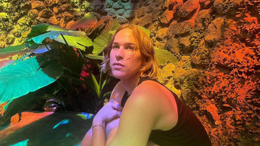 Tommy Dorfman assise en tailleur sous des vitraux arc-en-ciel, entourée de murs en pierre et de reflets colorés.