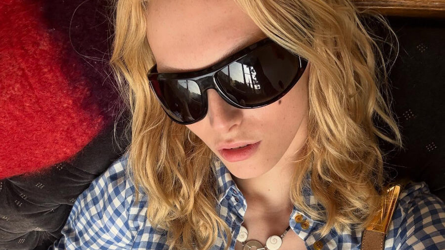 Alex Consani assise dans une pose détendue, portant une chemise vichy bleue, une mini-jupe blanche, des lunettes de soleil foncées et un sac à bandoulière.