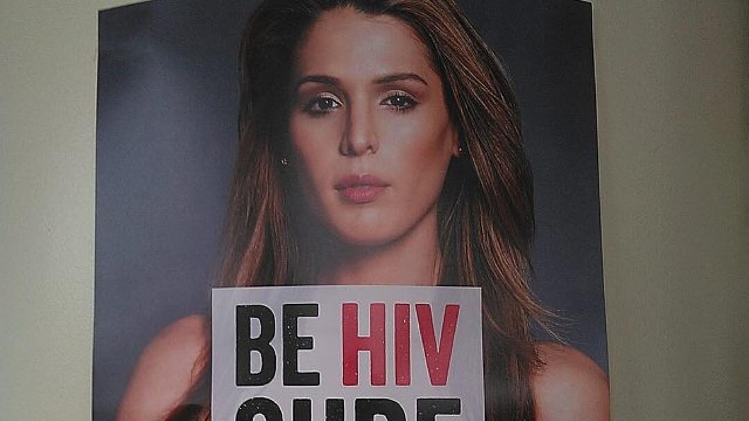 Carmen Carrera posant sur une affiche de sensibilisation au VIH, tenant une pancarte avec le texte “Be HIV Sure”.
