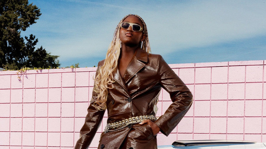 Zaya Wade pose en extérieur avec un manteau en cuir marron et des lunettes de soleil, à côté d’une voiture bleu clair lors d’un shooting mode.