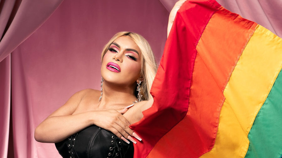 Wendy Guevara en robe corset noire, tenant un drapeau arc-en-ciel devant des rideaux roses pour MAC Pride 2024.