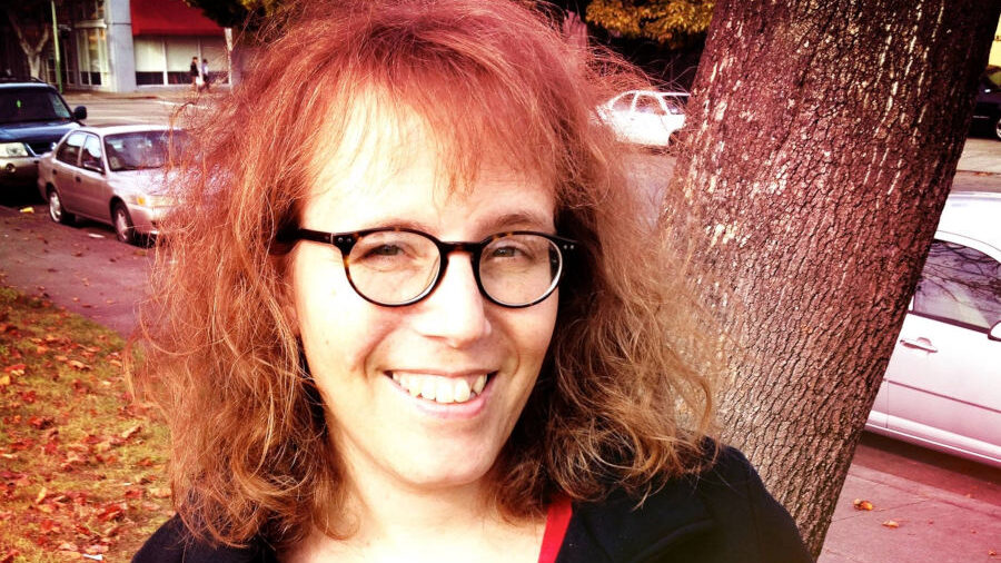 Julia Serano souriante en extérieur près d’un arbre, portant des lunettes et un manteau noir sur un haut rouge, avec des voitures garées derrière elle.