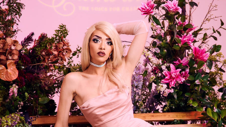 Arisce Wanzer portant une robe en satin rose, posant sur un banc en bois entouré de fleurs roses et de plantes décoratives.